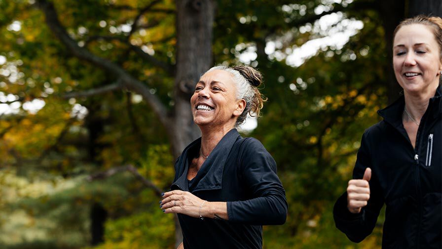 Duas mulheres a correr ao ar livre num parque de outono, sorridentes e vestidas com roupa desportiva.